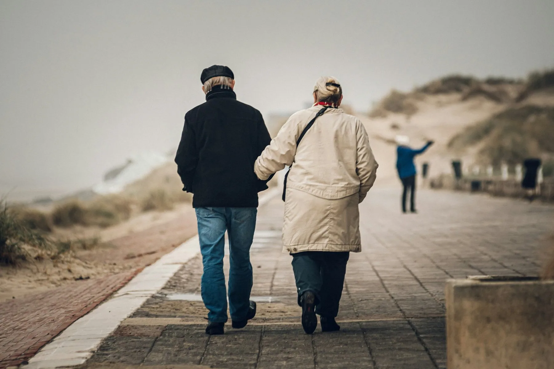 Eldre par som går arm i arm langs en strandpromenade, sett bakfra