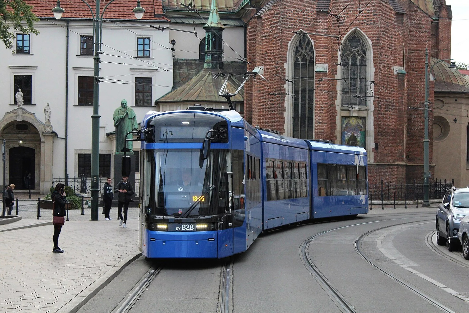 En blå Stadler Tango-spårvagn på linje 18 framför en gotisk kyrka i Kraków